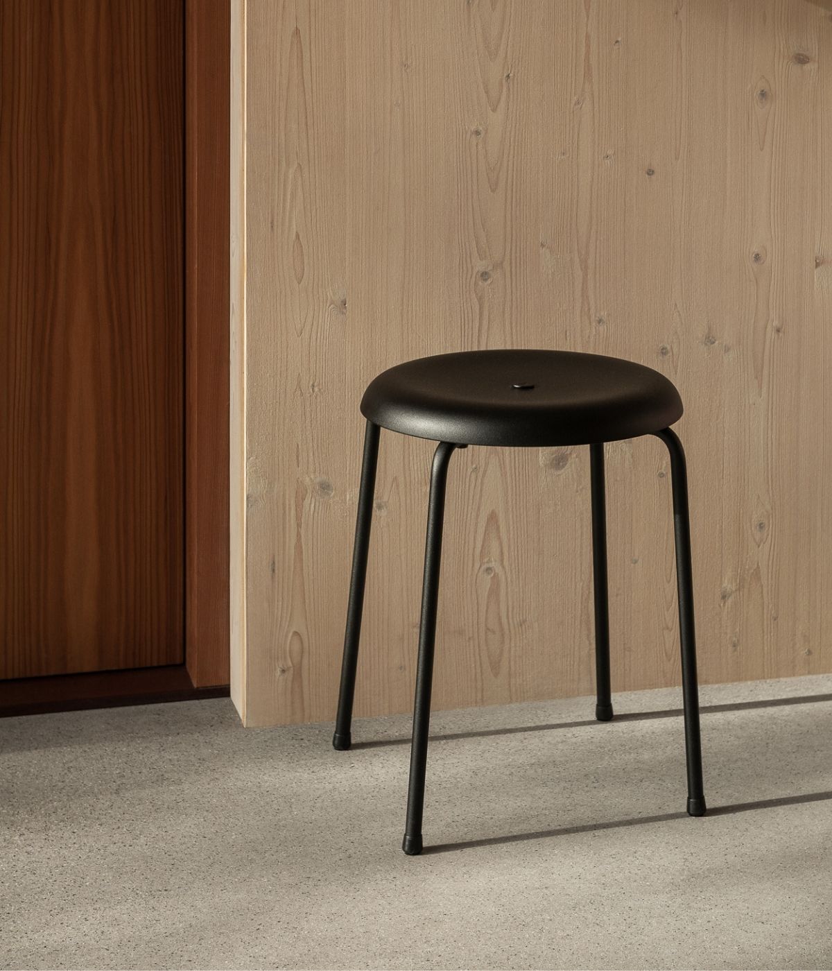 Taburett/Hocker von Lammhults in Schwarz vor einer Wand aus Holz. Das klare, moderne Design des Hockers wird durch die warme Holzstruktur im Hintergrund perfekt ergänzt, wodurch eine stilvolle und einladende Atmosphäre entsteht.“