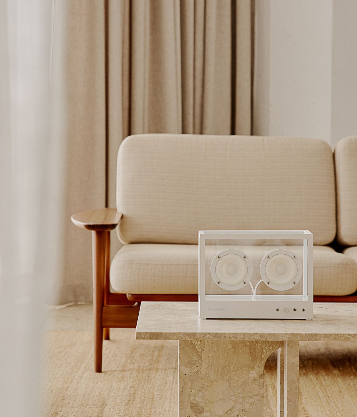 Small Transparent Speaker in Weiß, stilvoll inszeniert in einem modernen Wohnzimmer. Der Lautsprecher steht auf einem dezenten Möbelstück, umgeben von einem Interieur mit klaren Linien, neutralen Farben und hochwertiger Einrichtung. Große Fenster, dezente Dekorationen und sorgfältig ausgewählte Möbelstücke unterstreichen das minimalistische, zeitgemäße Ambiente. Das transparente Design des Speakers fügt sich harmonisch in die moderne Umgebung ein und betont seinen Anspruch als technisches wie ästhetisches 
