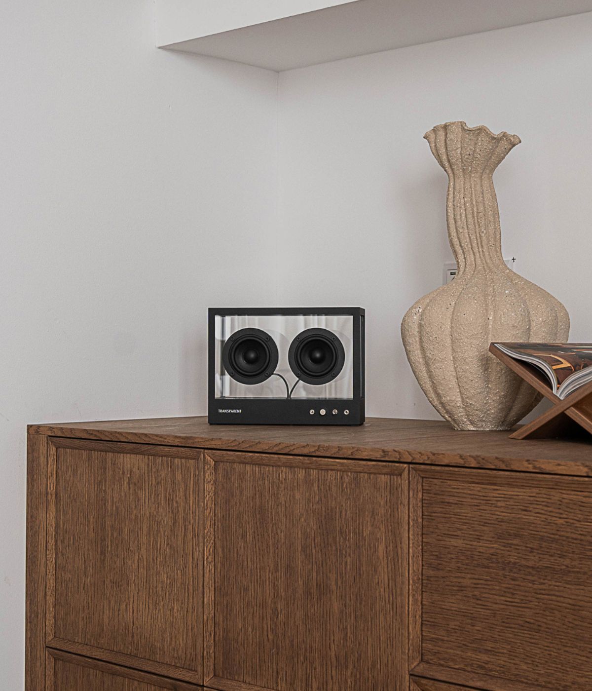 Small Transparent Speaker frontal aufgenommen, positioniert auf einem Holzschrank. Das klare, transparente Gehäuse zeigt direkt nach vorn, sodass die beiden Lautsprechertreiber sowie die interne Technik deutlich sichtbar sind. Das Gerät steht auf einer warmen, strukturierten Holzoberfläche, deren natürliche Maserung einen schönen Kontrast zum modernen, minimalistischen Design des Speakers bildet. Im Hintergrund sorgt eine dezente Rauminszenierung für eine wohnliche, stilvolle Atmosphäre, die das Produkt in 