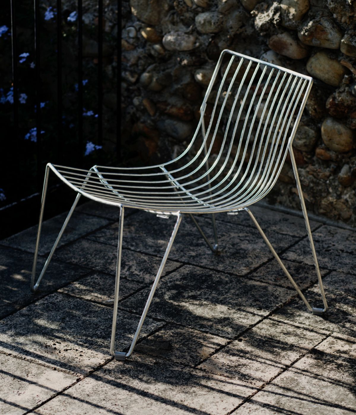TIO Easy Chair in galvanisierter Metalloptik von Massproductions, vor einer rustikalen Steinwand im Außenbereich auf gepflastertem Boden platziert – robuste, minimalistische Sitzgelegenheit, die modernen Industriedesign-Charme mit natürlicher Umgebung verbindet  
