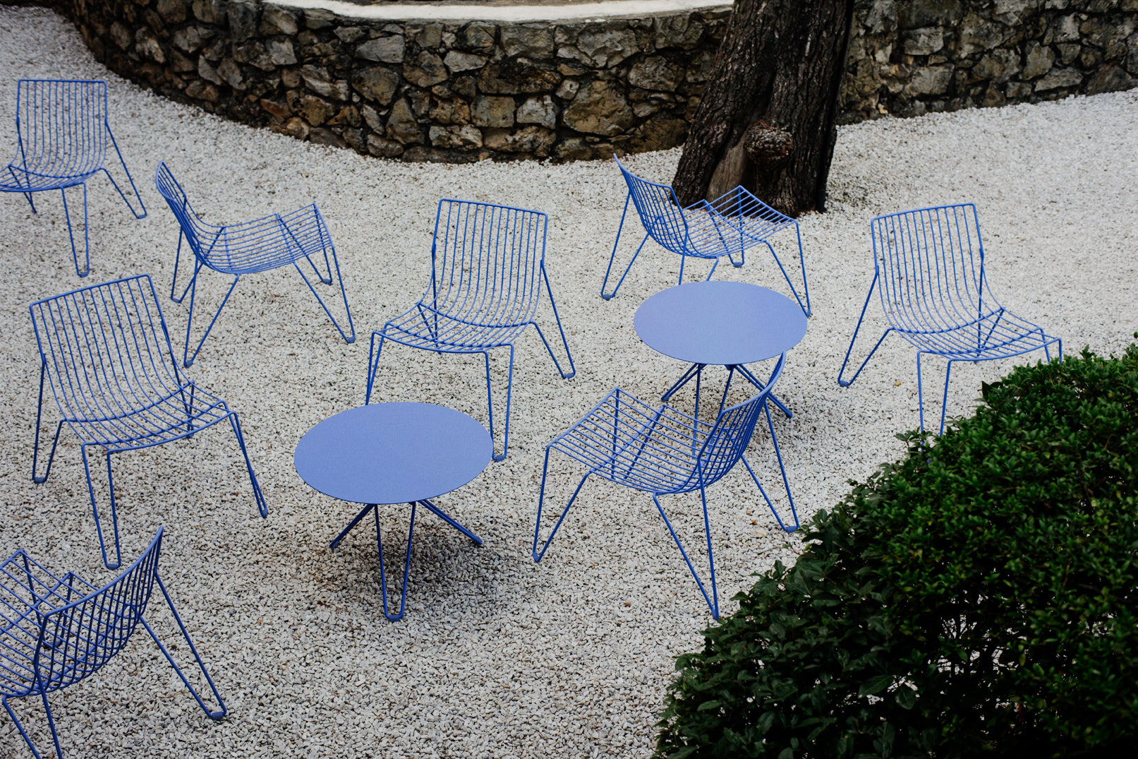 Mehrere TIO Easy Chairs in sanftem Blau von Massproductions, zusammen mit passenden blauen Tischen, im Kiesgarten mit Pool im Hintergrund inszeniert – eine harmonische Ansammlung von minimalistischen Möbeln, die das moderne Outdoor-Design mit der natürlichen, eleganten Atmosphäre des Gartens verbinden
