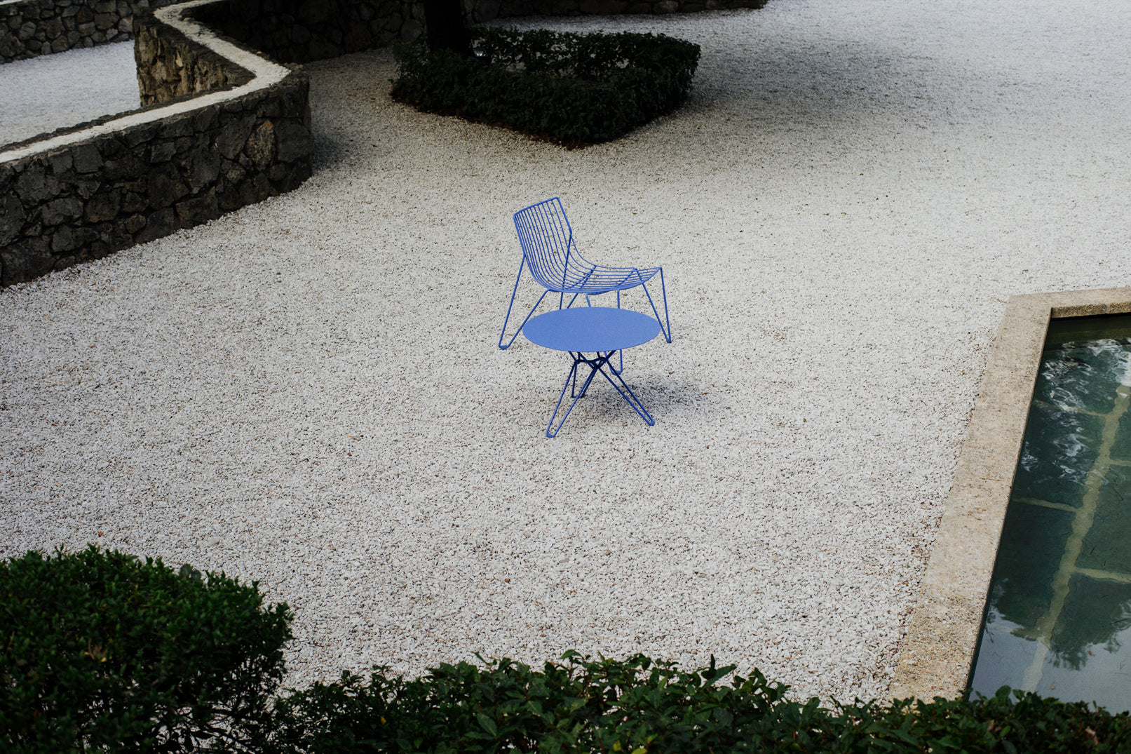 Zwei TIO Easy Chairs in sanftem Blau von Massproductions, aus leicht erhöhter Perspektive im Kiesgarten mit Pool im Hintergrund inszeniert – elegante, minimalistische Stühle, die perfekt mit der modernen, natürlichen Umgebung harmonieren und einen entspannenden, stilvollen Outdoor-Bereich schaffen

