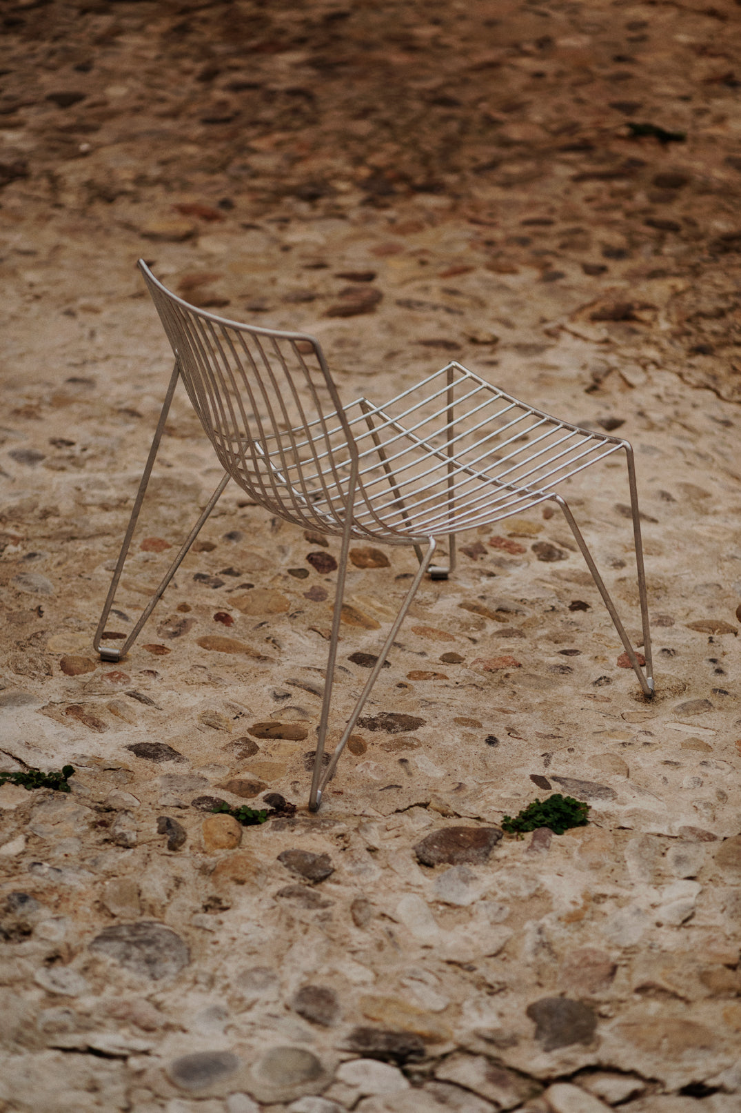 TIO Easy Chair in galvanisierter Metalloptik von Massproductions, frontal auf Steinboden platziert – moderner Outdoor-Stuhl mit filigraner Gitterstruktur und industriellem Charakter, ideal inszeniert in einer natürlichen Umgebung
